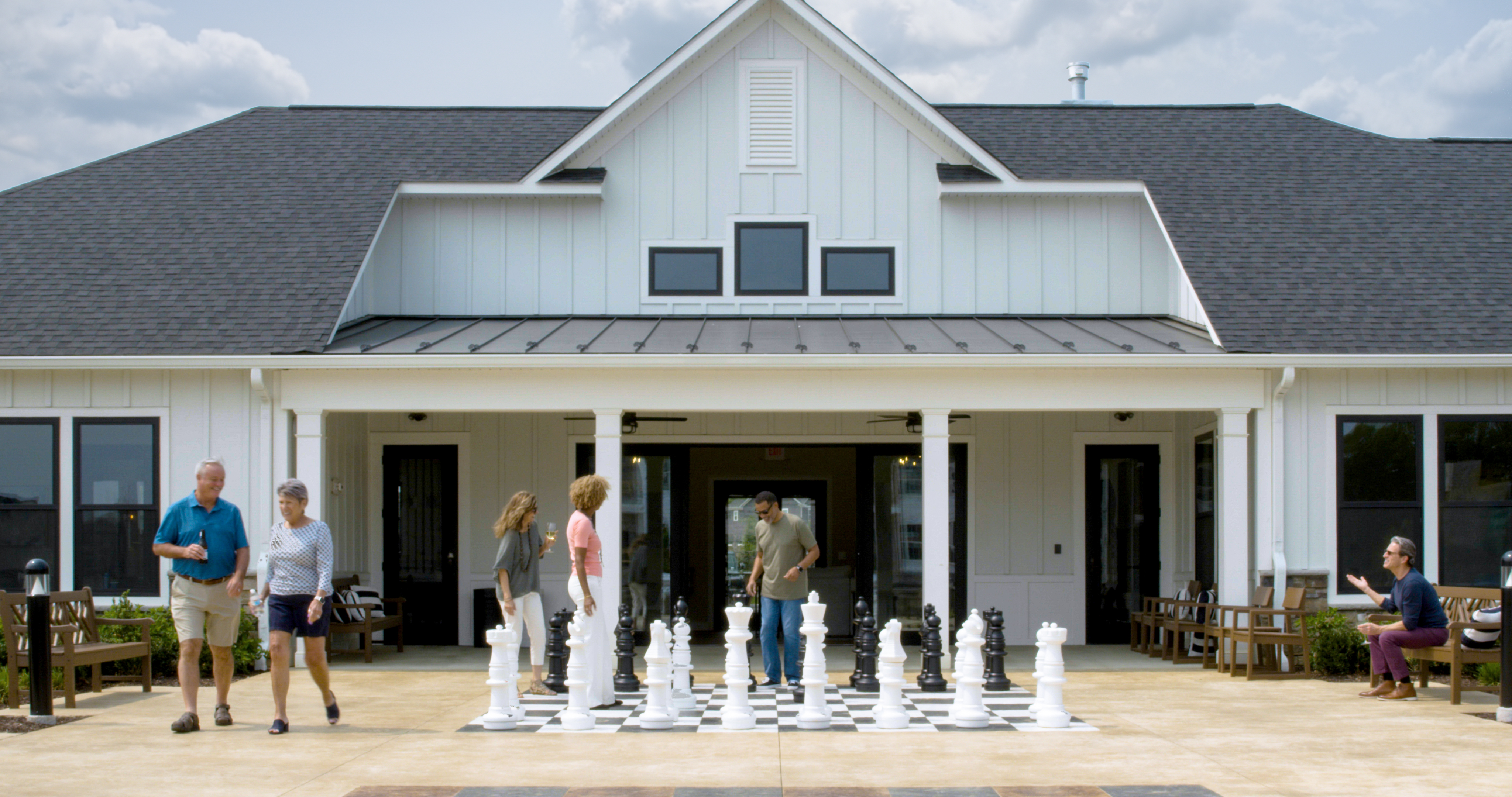 Adults gather outside a clubhouse where two people play a large outdoor chess game.