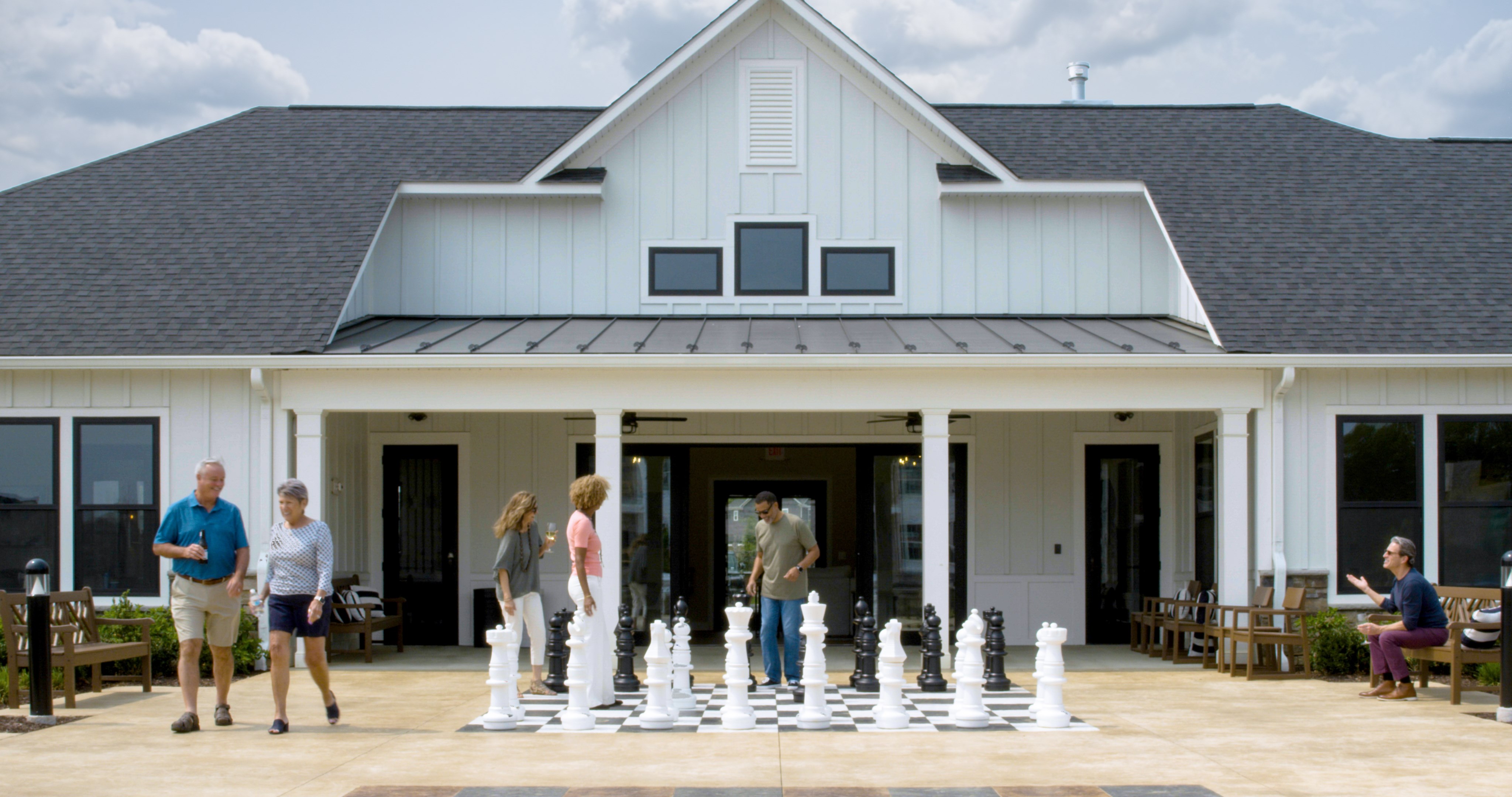 Adults gather outside a clubhouse where two people play a large outdoor chess game.
