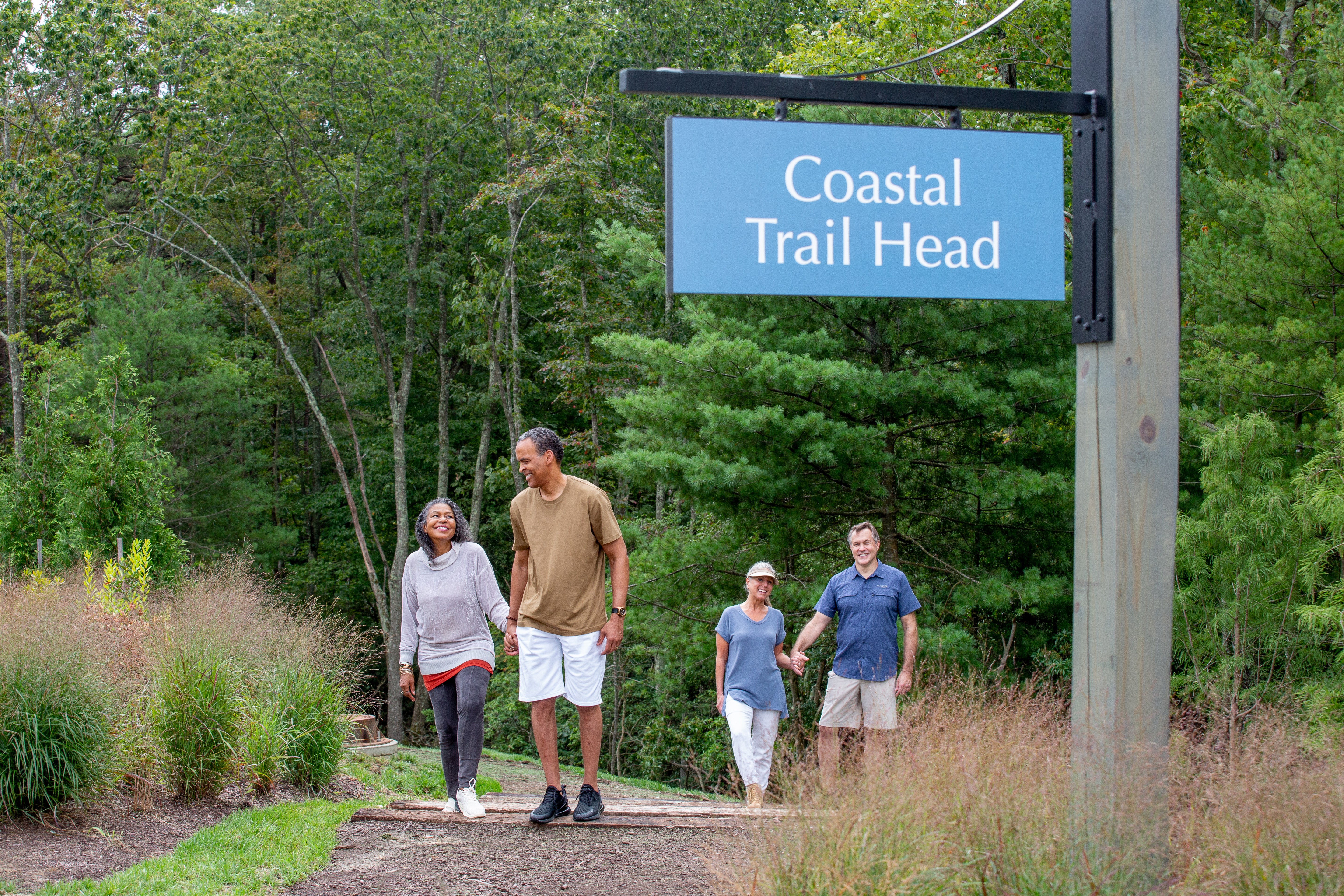 The image shows four people walking near a sign that reads “Coastal Trail Head” at Embrey Mill, a scenic wooded area with walking trails.