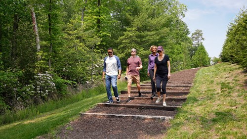 A group of adults walks down a wooded trail in Embrey Mill, a master-planned community in Stafford, VA. Surrounded by lush greenery and well-maintained paths, the scene reflects the community’s active lifestyle and connection to nature.