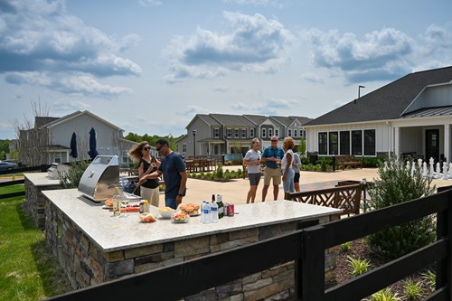 People socializing and grilling food at an outdoor community barbecue area near modern houses.