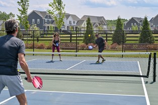 Residents playing a game of pickleball on an outdoor court in Stafford, VA, with modern homes and landscaped greenery in the background at Embrey Mill.
